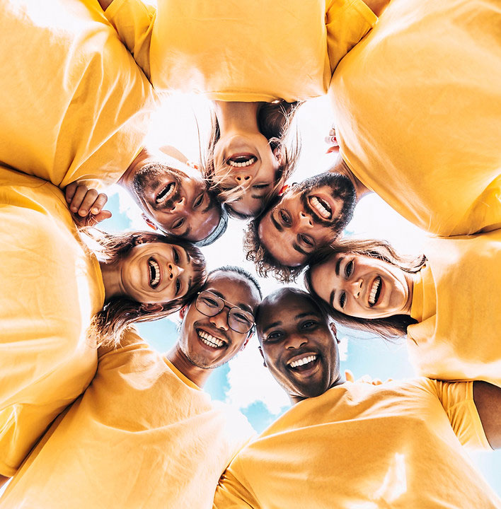 The image shows a group of seven individuals smiling at the camera, with their arms outstretched forming a circle around each other. They are wearing yellow shirts, suggesting they might be part of an organized team or group.