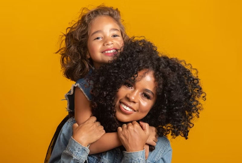 A woman with curly hair hugs a young child, both smiling against a yellow background.