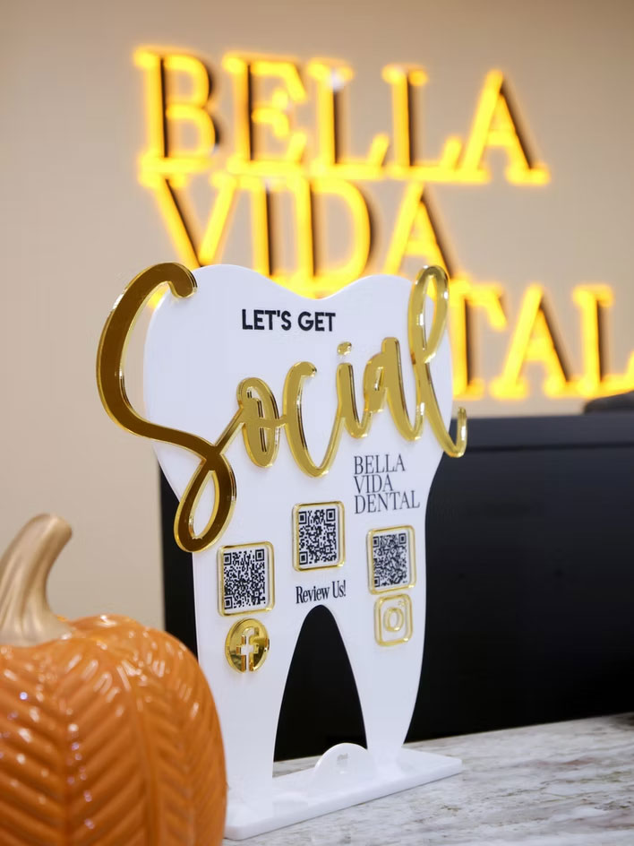 The image features a dental office sign with a tooth-shaped sign that reads  Bella Vida Dental  and a social media QR code, placed on a desk with a Halloween pumpkin.