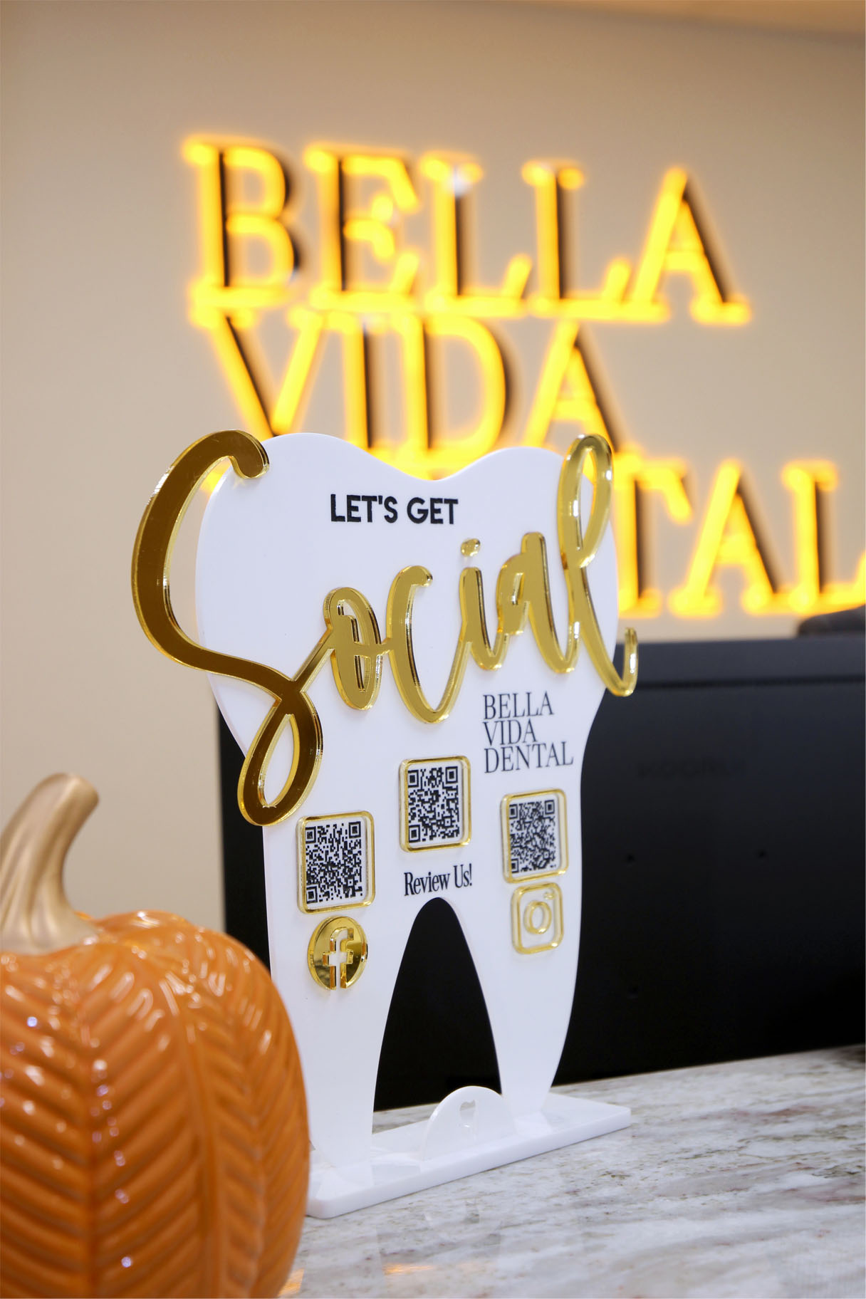 The image shows a sign with the name  Bella Vida Social  accompanied by a toothbrush-shaped sign that says  Let s get social  and a QR code, placed on a countertop next to a pumpkin.