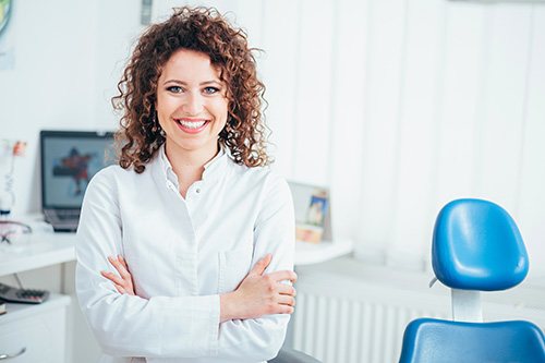 The image shows a woman standing in an office environment, smiling at the camera, with her hands crossed over her chest. She has curly hair, is wearing a white shirt, and is positioned in front of a desk with a computer monitor and chair.