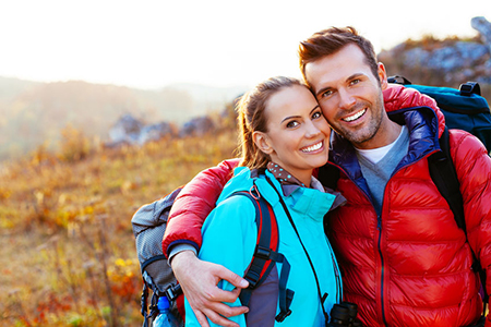 A man and woman are embracing outdoors, both wearing backpacks and smiling, with a scenic landscape featuring autumn foliage behind them.