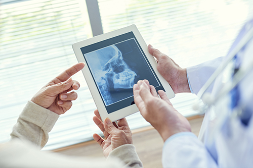 The image shows two individuals interacting with a tablet screen displaying an X-ray image while standing in a medical setting, likely a hospital or clinic.