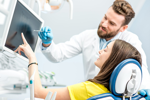 A man in a white coat stands next to a woman seated on a dental chair, both looking at a computer screen  they are in a dental office setting.