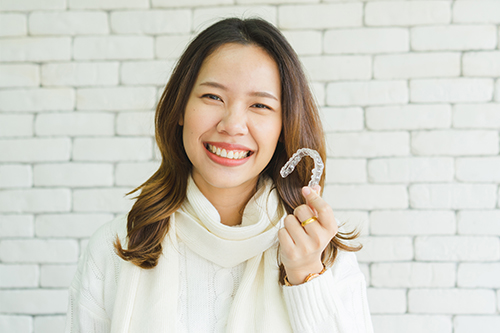 A woman holding a white smiley face with her left hand against a brick wall background.