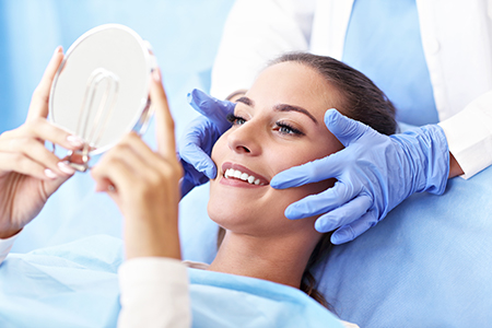 In the image, a woman is lying on a dental chair with her eyes closed while a dentist is examining her teeth with a mirror, wearing blue gloves and a white coat.