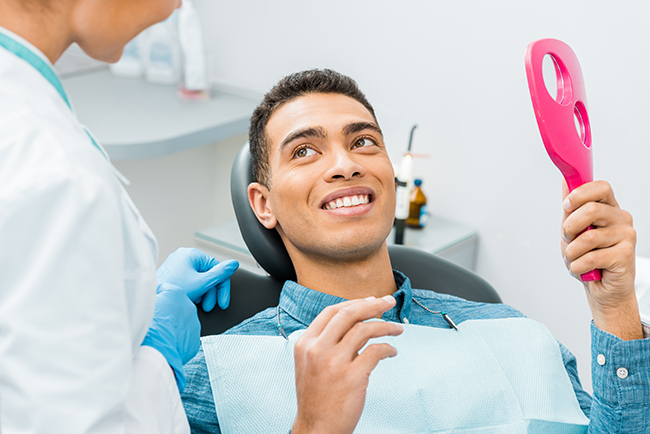 A man is seated in a dental chair while holding an electric toothbrush, with a dental hygienist standing behind him, both smiling. They are in a dental office setting.