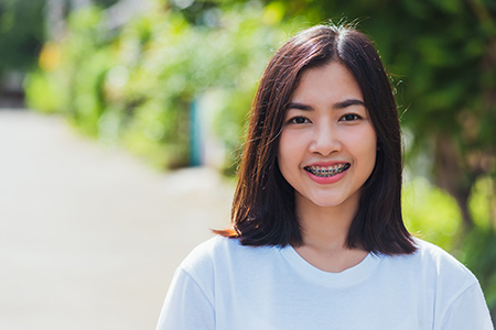 The image shows a young woman smiling at the camera with short hair, wearing a white shirt and standing outdoors against a blurred background of greenery.