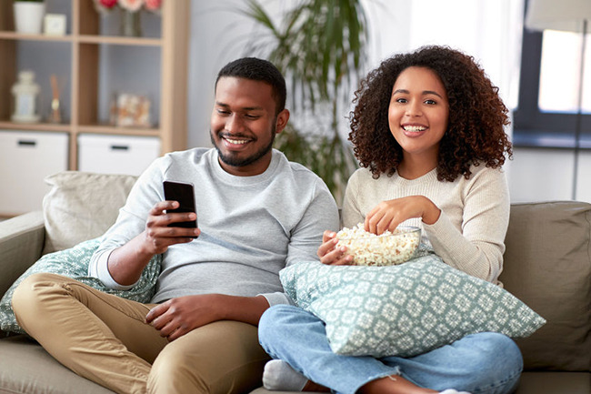 The image shows a man and woman sitting on a couch, enjoying popcorn while watching television.